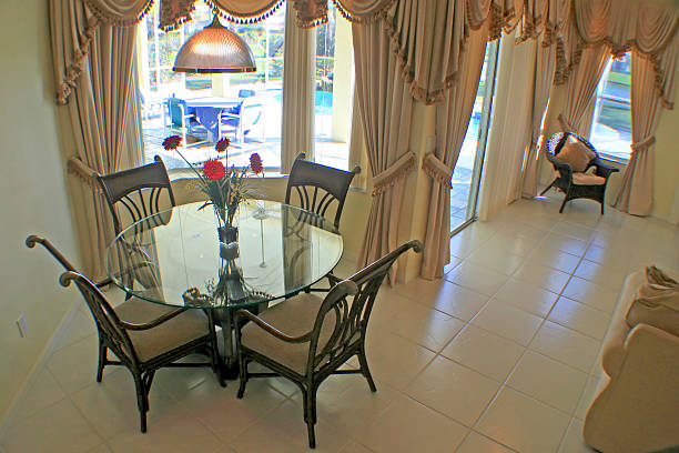 Стеклянный обеденный стол с четырьмя стульями на хорошо освещенной, элегантной кухне с большими окнами и задрапированными шторами.